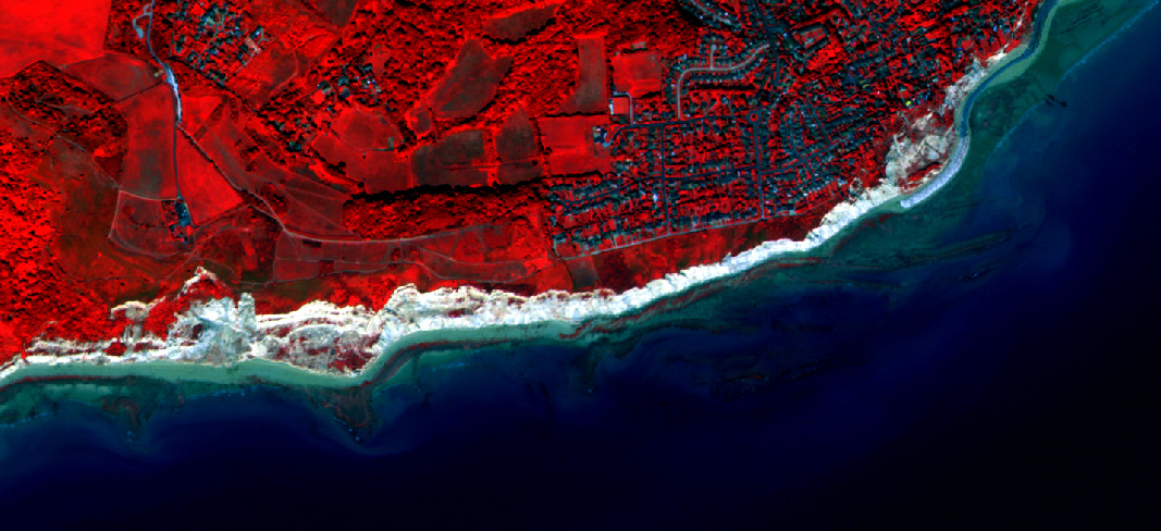This is a false-color satellite image depicting a coastal landscape. Healthy vegetation, including forests and agricultural fields, appears in shades of bright to dark red, indicating high reflectance in the Near-Infrared spectrum. Residential areas and urban infrastructure, characterized by buildings and roads, are visible as darker, mottled regions embedded within the vegetated areas. A distinct white to light grey band outlines the coastline, suggesting exposed rocky cliffs or sandy beaches. The ocean or sea appears in dark blue, with lighter blue and greenish hues in the shallower coastal waters, indicative of bathymetry, submerged features like seagrass, or sediment. The image shows a detailed view, allowing individual buildings, field patterns, and coastal erosion features to be discerned.