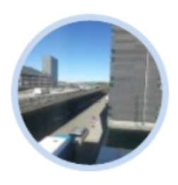 A circular, blurred aerial photograph depicting an urban or suburban landscape. The central feature is a wide, multi-lane road, possibly a highway, with several indistinct vehicles visible. On both sides of the road, there are tall buildings, suggesting an urban environment. The sky above is clear blue. The image is contained within a light blue circular border. Due to the heavy blurring, specific details of the buildings, vehicles, or surrounding terrain are not discernible.