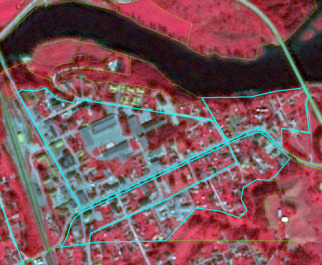 This map displays a False Colour Infrared (FCIR) satellite or aerial image of an urban area bordered by a dark blue body of water, likely a river, at the top. Healthy vegetation appears bright red, prominently along the riverbanks and surrounding the settlement. Impervious surfaces such as buildings and roads in the urban core are depicted in shades of grey and white. A prominent linear feature, likely a road or railway, runs diagonally through the western part of the urban area. Overlaid on the imagery are two sets of vector lines: light blue polygons delineate specific urban blocks or developed zones within the settlement, and thin green lines outline larger, irregular land parcels, including areas along the river and encompassing the broader urban and vegetated landscape. No scale bar, compass, legend, or reference year is visible.