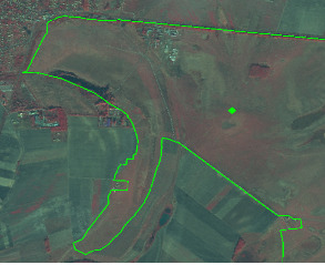 False-colour infrared (FCIR) satellite or aerial imagery displaying a mixed rural and peri-urban landscape. The imagery uses reddish tones for vegetation (darker red for denser vegetation, lighter red/pink for sparser areas) and grey/blue tones for non-vegetated or artificial surfaces. A large, irregular area is prominently delineated by a bright green polygon boundary. Within this delineated area, a single bright green circular point marker is visible towards the upper right. The landscape within and around the outlined area features distinct rectangular agricultural fields, patches of natural or semi-natural vegetation, and some greyish areas that may represent artificial surfaces or bare ground. No scale, compass, legend, or specific geographic location (country, region) is visible.