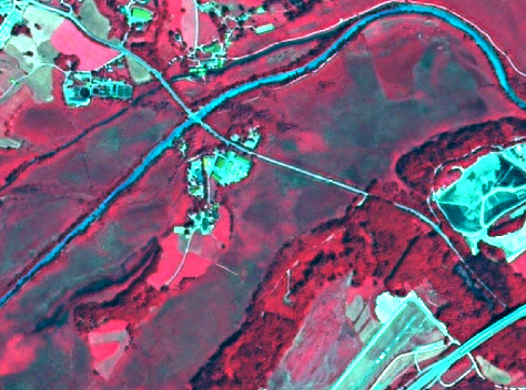 False-colour aerial or satellite imagery depicting a landscape with diverse land cover and land use. Vegetation, including forests and tree lines, is represented in various shades of dark red and magenta. A prominent river or stream flows from the upper left to the lower right, appearing bright cyan/light blue, with a white line along its banks, likely indicating a riparian zone or bare earth. Agricultural fields or bare soil areas are visible as lighter pinkish-purple or greyish-white patches, some with distinct rectangular shapes. Built-up areas and roads appear white or light grey, with a main road crossing the river via a bridge in the central portion of the image. In the upper-left and lower-right, a larger road network, possibly a highway, is visible. Specific features include: circular structures (possibly water treatment tanks or industrial ponds) coloured white/light blue near the centre-left; multiple light green rectangular areas, likely sports fields, surrounded by buildings near the centre-right; and a larger complex of light blue water bodies or industrial ponds in the far right. The image displays a mix of natural riparian and forest areas, cultivated land, and developed infrastructure.