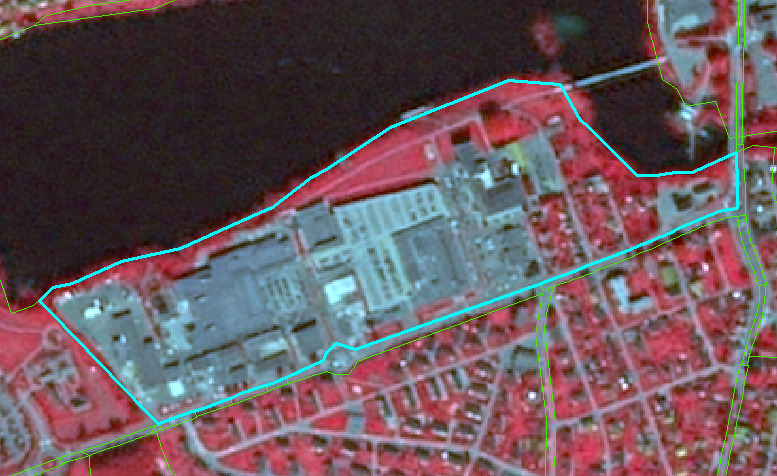 This is a false-colour infrared aerial image depicting a mixed landscape with an industrial complex, a large water body, forested areas, and residential zones. The image uses infrared bands where healthy vegetation appears bright red/pink, water bodies appear dark blue/black, and impervious surfaces like buildings and roads appear light blue/grey. A large industrial complex, consisting of numerous light blue/grey buildings and associated infrastructure, is centrally located and clearly delineated by a light blue (cyan) polygon boundary. To the north and northwest of this complex is a dark blue/black water body (likely a lake or large pond). Adjacent to the water and the industrial complex, especially to its north, are areas of dense vegetation shown in bright red, indicating a forest or heavily vegetated land. South and east of the industrial complex, the landscape transitions into residential areas, characterised by smaller, reddish-pink building rooftops and a network of roads (thin green lines overlaid on the image, potentially representing cadastral or road network data). Additional green lines are visible, outlining the water body and defining other land parcels. No scale bar, compass, legend, or reference year is visible.