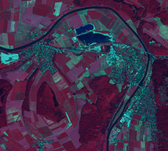 A false-color infrared satellite or aerial image depicts a diverse European landscape. A prominent, wide, meandering river, appearing dark blue/cyan, flows from the top-left, forms a large curve around a central landmass, and exits towards the bottom-right. Within this river bend, two distinct, interconnected, darker blue water bodies, likely reservoirs or natural lakes (potentially mapped under CLMS category *8.2.1.0 Natural lakes*), are visible. The surrounding land cover is dominated by irregularly shaped agricultural fields, displayed in shades of reddish-pink, indicating vegetated crops or bare soil. Patches of denser, dark red/maroon vegetation, representing forests or woodlands, are scattered across the scene, notably in the lower right. Settlements or urbanized areas appear as lighter blue-green/cyan tones with irregular patterns and clustered structures, particularly along the inner riverbanks. The false-color infrared palette highlights vegetation as red, water as dark blue, and built-up areas with cyan tones, typical for remote sensing applications in land cover analysis.