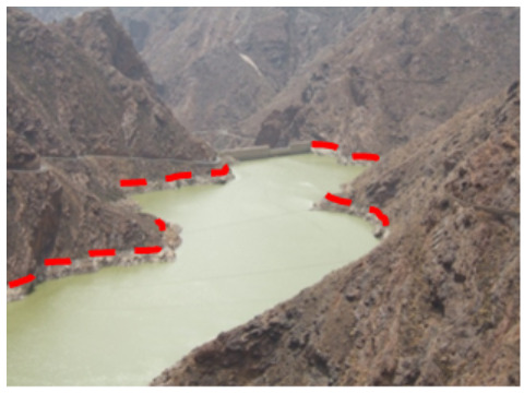A photograph depicting a reservoir situated in a mountainous, arid landscape, likely in the Canary Islands, Spain. A concrete dam structure is visible across the water body in the background. The current water level along the rocky shoreline is highlighted by a red dashed line, which appears to correctly delineate the water body's limits. The surrounding context indicates this image demonstrates the 'correct interpretation' for delimiting water bodies using the current water level for mapping purposes, particularly in the context of temporal fluctuations. The image credit is M. Palacios.