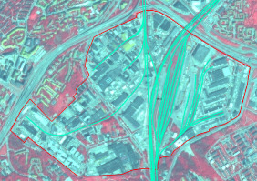 This is an aerial image of an unlabelled urban or industrial area, displaying land cover in an infrared representation, where vegetated areas appear in red hues and built-up structures or impervious surfaces appear in grey and white tones. A red polygon outline defines a specific zone within this area. Within and around this outlined zone, aqua-coloured linear features represent extensive transportation infrastructure, including a complex of multiple parallel railway tracks or roads dominating the central-right part of the image, and other connecting lines. The red-outlined zone encloses a mix of industrial or commercial buildings and significant transportation infrastructure. No scale bar, compass, legend, or reference year is visible in the image.