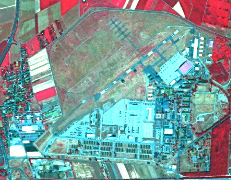 A false-color satellite or aerial image depicting a diverse landscape composed of urban, industrial, and agricultural features. The image uses a colour scheme where healthy vegetation appears bright red, impervious surfaces (such as buildings, roads, and bare soil/concrete) are light blue or cyan, and areas with sparse vegetation, dry grass, or fallow land appear greenish-brown or tan. Key features visible include: - A prominent complex of light blue/cyan buildings, including several large rectangular structures and numerous smaller ones arranged in rows, indicative of an industrial facility, large campus, or military base, located in the central and right portions of the image. - A large, irregular-shaped open area with greenish-brown hues, possibly an airfield or testing ground, occupying the upper central portion. This area contains a curved track and some small, regularly spaced dark features. - A network of roads and parking lots (light blue/cyan) connects the various structures within the industrial/urban complex. - To the left, a residential-like area with smaller light blue/cyan buildings is visible, bordered by distinct rectangular agricultural fields, some appearing bright red (healthy vegetation) and others bright white/yellow (bare soil or harvested fields). - The periphery of the image, particularly the top, left, and bottom, shows additional agricultural fields and areas of dense red vegetation, likely forests or active crop fields. A road network with what appears to be an interchange is visible in the top-left corner. No scale bar, compass, or explicit date is visible on the image.
