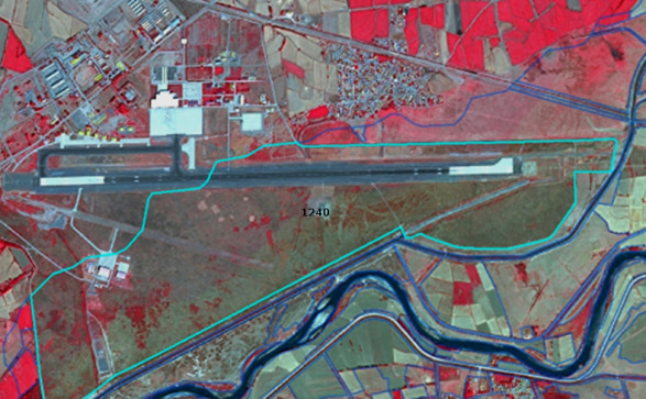 This map displays a false-colour infrared satellite image of an airport and its surrounding land cover. The airport infrastructure, including its main runway, a secondary shorter runway, buildings, and associated tarmac, appears in shades of grey, white, and light blue. Extensive vegetated areas within and adjacent to the airport, likely grassland or agricultural fields, are rendered in shades of red, characteristic of vegetation in false-colour infrared imagery. A light blue polygon boundary encloses the core airport area, and within this boundary, the numerical label '1240' is visible, which corresponds to the CORINE Land Cover (CLC) class code for 'Airports'. A river, appearing dark blue, forms the southern border of the delineated airport area. Beyond the main airport boundary, the landscape comprises agricultural fields (shown in red and brown hues) segmented by thin dark blue lines indicating parcel divisions, and a small settlement with grey-toned buildings is visible to the northeast. No scale bar, compass rose, or explicit date information is present within the image.
