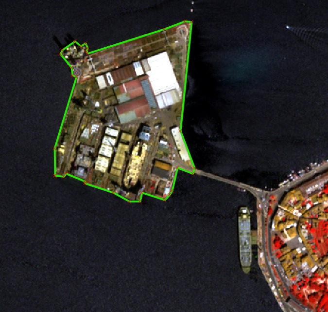 An aerial or satellite map displays a coastal industrial area, likely a port facility, bordered by a large body of dark blue water. The main industrial landmass is delineated by a bright green polygon, with small red dots marking its vertices. Within this polygon, numerous buildings are visible, characterised by white, grey, brown, and reddish roofs, indicative of various industrial or commercial structures. A long, narrow bridge or causeway connects the industrial area to an adjacent, densely built-up urban area visible on the right side of the image. Buildings in this urban area appear in red and yellow tones, suggesting a false-colour rendering. A large, grey vessel, possibly a cargo ship or barge, is present in the water near the bridge. No scale bar, compass orientation, legend, or data source/reference year is provided.
