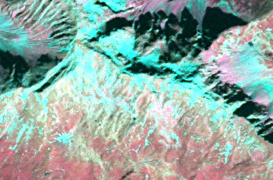 A false-colour satellite or aerial image depicting a rugged mountainous landscape. The image shows a complex terrain with pronounced ridges and valleys, particularly in the upper left and central-upper regions, exhibiting varied textures and strong relief. Numerous small to medium-sized water bodies appear in bright turquoise/cyan, scattered across the lower half and upper right of the image. The dominant land cover in the central and lower areas is rendered in reddish, pinkish, and purplish tones, characteristic of bare soil, sparse vegetation, or exposed rock formations in false-colour infrared imagery. Darker areas, especially in the upper central region, suggest shadows or specific geological features. No scale, orientation, or temporal context is visible.