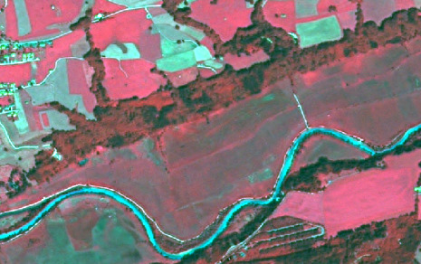 False-colour infrared satellite imagery of a rural landscape, likely showing land cover and land use. The image depicts a meandering river rendered in bright cyan/turquoise, with a white line closely delineating its banks. Dense linear vegetation, appearing in deep red/brown, follows the river and forms boundaries around large agricultural fields. These fields are visible in varying shades of reddish-pink and light green/turquoise, indicating different crop types, growth stages, or bare soil. A small settlement or urban area, characterized by clusters of light blue/cyan features with white outlines, is located on the far left of the image. A distinct linear bright cyan feature crosses the river, suggesting a bridge or road. The colour scheme is typical of false-colour composites, where healthy vegetation is represented by strong reds, water by cyan/blue, and impervious surfaces or bare ground by lighter colours.
