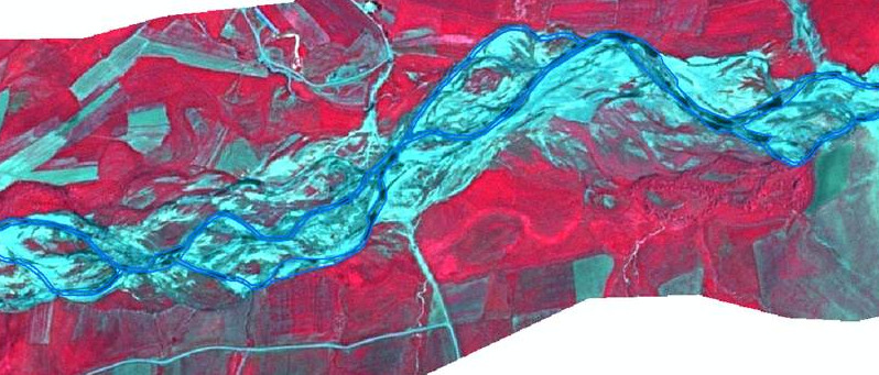 A false-colour infrared aerial or satellite image displaying a winding, braided river system outlined in blue, surrounded by varied land cover. The river channel and its immediate floodplain appear in light blue/cyan, indicating water or wet and sandy areas. A prominent blue line delineates the boundary of the main river corridor, potentially representing a riparian zone or active floodplain. Surrounding the river, the landscape shows large areas of vibrant red, characteristic of dense vegetation such as healthy forests or crops. Interspersed with these red areas are greenish-blue rectangular patches, likely representing agricultural fields or other cultivated land with different vegetation characteristics or soil moisture. Other areas appear as reddish-brown, indicating sparser vegetation or bare soil. No scale bar, compass orientation, legend, or data source/year is visible.