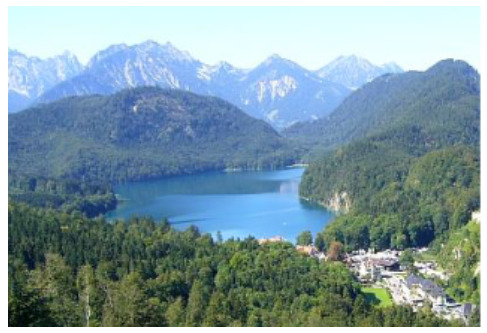 A wide-angle landscape photograph features a large, deep blue lake as the central element, reflecting the clear sky. The lake is surrounded by densely forested hills and mountains, primarily covered with dark green coniferous trees. In the background, rugged, snow-capped or bare rock mountains rise prominently under a clear, bright blue sky. On the lower right side of the image, nestled between the forest and the lake, a small village or settlement is visible, with several light-coloured buildings and some open green spaces. The overall scene depicts a natural, mountainous environment with minimal human development concentrated in one small area. No text is visible within the image.