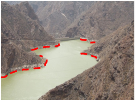 This photograph depicts a reservoir nestled within a steep, rocky, mountainous landscape, identified as being in the Canary Islands, Spain. The water in the reservoir appears a pale greenish-yellow. A dam wall is visible in the background. A red dashed line delineates the current visible water level along the shore. According to the accompanying text, this image illustrates the 'wrong interpretation' for the delimitation of water bodies, specifically in the context of using the maximum water level for mapping, implying that the depicted water level is not the maximum. Credit: M. Palacios.