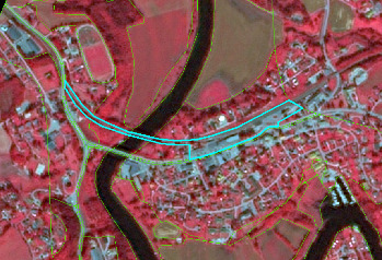 This map presents a false-colour infrared aerial or satellite image of a river valley. The main river meanders through the scene, appearing in dark blue/black. Healthy vegetation, such as forests and agricultural areas, is highly reflective in the infrared spectrum and appears in shades of dark red/pink. Urban infrastructure, including roads and buildings, is visible in lighter grey and white tones. Green lines delineate various land cover parcels or administrative boundaries across the landscape. A prominent cyan-coloured polygon highlights an elongated urban and infrastructure strip located on the right bank, running parallel to the river and a main road. Other identifiable features include a sports complex with a running track and field on the left bank of the river and a potential harbour or marina in the bottom right corner. No scale bar, compass orientation, legend, data source, or reference year is visible in the image.