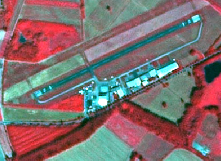 This is a false-colour infrared (NIR) satellite image depicting an airfield and its surrounding rural landscape. The image uses a colour composite where healthy vegetation, such as crops and forested patches, appears bright red. Areas of bare soil, fallow fields, or less vigorous vegetation are shown in shades of green and beige. A small dark blue pond, characteristic of water bodies in NIR imagery, is visible in the upper-left quadrant. The central feature is an airfield, comprising at least one main runway and parallel taxiways, which appear grey to light blue. A cluster of white to light blue buildings, likely hangars and support facilities, is located adjacent to the runways. Linear features resembling roads or tracks traverse the agricultural fields and connect to the airfield complex.
