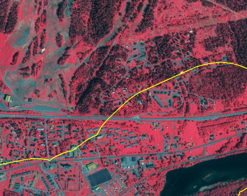 False-colour infrared satellite image of Uleforss, Norway, captured by SPOT-5 at 2.5m resolution on 2010-08-11, using a 1/2/3 (Green, Red, Near-infrared) band combination. The image depicts a diverse landscape including: dense vegetation (forests) appearing in deep red, particularly in the upper and lower sections; a major linear transport infrastructure (road/highway) running horizontally across the middle, shown in grey/blue; and an adjacent settlement with clusters of buildings in light grey/blue. North of the main road, a large area of lighter red/pink vegetation with distinct patterns is visible, identified as a golf course based on external context, interspersed with scattered smaller buildings (possibly holiday homes or cabins) within the forested landscape. A dark blue water body, likely a lake or wide river, is present in the bottom right corner. A prominent bright yellow line delineates a boundary, starting from the lower left, following along the main road, and then curving northwards to encompass portions of the forested area and the golf course. This line likely indicates a land cover classification boundary. Source: CNES 2011©, Distribution Airbus DS/Spot Image.
